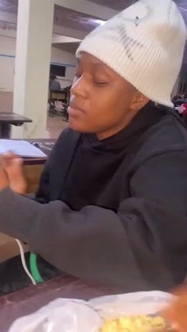 A young black person wearing a white beanie and a black hoodie is sitting at a wooden table, looking to their left with their eyes closed and a slight smile. Their right hand is raised to their face, with their index and middle fingers extended in a peace sign. In the background, there are other people sitting at tables, and a green bottle is visible on a table. The lighting is dim.
