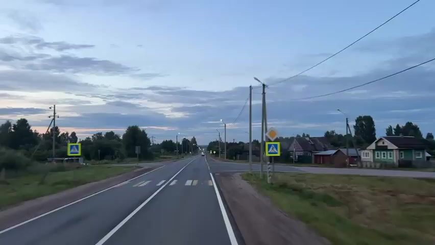 The video shows a view from a moving vehicle driving down a two-lane road in a rural area at dusk. The sky is overcast with scattered clouds, and the ambient light suggests it is either early morning or late evening. Streetlights are visible along the road and are illuminated. On both sides of the road, there are trees, grassy areas, and houses typical of a village. Road signs are visible, including pedestrian crossing signs and diamond-shaped warning signs. The road has clear lane markings, including a solid white line on the right edge and dashed white lines separating the lanes. A few cars are visible on the road, including one in the distance with its headlights on. The vehicle appears to be moving at a moderate speed.