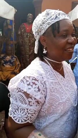 A woman in a white lace top and white head wrap is seen holding a framed picture of a man in a red outfit. She is smiling and appears to be speaking to someone off-camera. Money is pinned to her clothing. Behind her, several people are gathered, some seated and others standing. The scene takes place outdoors, likely in a courtyard or outdoor gathering area, with buildings visible in the background.