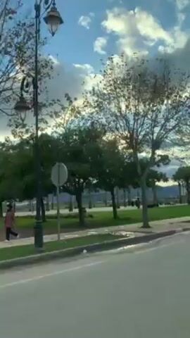 A car is driving along a paved road adjacent to a park. The park has green grass, trees, and a fenced-in athletic court with a Moroccan flag attached. Some people are walking in the park.  The sky is partly cloudy with a lot of light reflecting off the clouds. There are light posts along the road with decorative globes. In the distance are hills or mountains. The road is gray and has a white stripe marking lanes. A parked gray car is near the court. The shot is taken from inside a moving vehicle.
