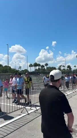 The video shows an outdoor scene with a clear blue sky and scattered clouds. Several people are walking towards a set of metal barriers, which appear to be set up to control crowd flow. Some individuals are wearing sports jerseys and carrying flags, suggesting they are attending an event. Palm trees and other foliage are visible in the background, along with some buildings and light poles. The ground is paved, and the overall impression is of a sunny day at an outdoor venue.
