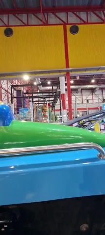 The camera is mounted on a blue and green ride, likely a small rollercoaster, in what appears to be a large indoor amusement park. The background features various structures including a jungle gym-like climbing structure, other rides, and open spaces with people walking around. The park's ceiling is visible, consisting of exposed beams and a yellow and red covering. The ride moves forward, creating a slight bump effect.
