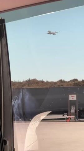 The video shows an airplane flying in the sky from the perspective of someone inside a vehicle. The airplane is small in the frame, suggesting it is at a distance. Below the airplane, a large truck is parked. The truck appears to be a tanker truck, with a large cylindrical tank. The truck has a sign on its side that reads "Panin Bank". There is also a gas pump visible next to the truck. The scene is an outdoor setting, likely a rest stop or a service area. The sky is clear and blue. There are trees in the background, suggesting a rural or suburban environment. The video is short and does not contain any dialogue or audible sound.