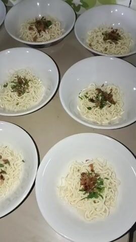 The video shows a top-down view of six white bowls filled with noodles, each topped with chopped green herbs and fried shallots. The bowls are arranged in a staggered pattern on a brown, speckled surface, possibly a table. The lighting suggests an indoor setting, possibly a kitchen or dining area. There are no people visible in the frame, and no actions or interactions are depicted. The noodles appear to be pre-cooked and portioned, ready to be served or further prepared.