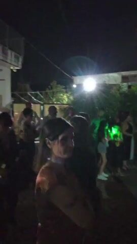 The video shows a nighttime outdoor gathering, possibly a party. Several people are visible, some in the foreground and others in the background. There are trees and bushes present, suggesting a garden or yard setting. In the background, a building or structure is partially visible, illuminated by bright lights. There's also a string of lights hanging above, and what appears to be a table with some decorations or items on it, including a prominent "18" sign made of lights. The overall atmosphere seems festive, with people interacting and what sounds like music playing.