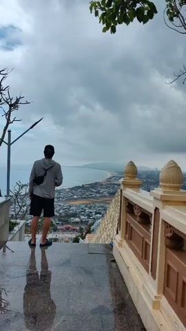 A man is standing on the steps of a high vantage point, possibly a temple or shrine, overlooking a coastal city. The man is wearing a light gray hooded jacket, black shorts, and a black baseball cap. He has a black bag slung across his shoulder and is wearing sunglasses. The sky is overcast, and the view extends to a beach and the ocean. In the distance, there are wind turbines along the coast. The stairs are marble, and there is a decorative railing beside them. The man looks around and appears to be admiring the view, holding a small electronic device in his hand. A bare tree stands next to the staircase.