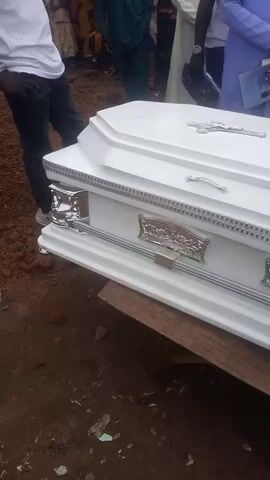 The video shows a white coffin with silver embellishments and a cross on top. It is placed on a wooden surface outdoors, with people gathered around. Some people are wearing white shirts and others are in traditional African attire. The ground appears to be dirt and there are some scattered leaves and debris. The mood seems somber.