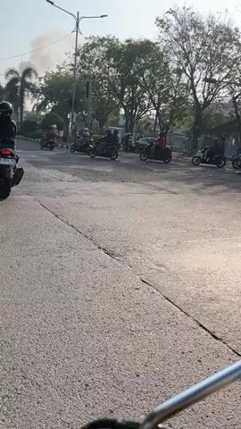 The video shows a group of motorcyclists riding on a paved road. There are several motorcycles, each with a rider, in the shot. Some riders are wearing helmets, while others are not. The environment appears to be an urban or suburban setting, with trees and buildings visible in the background. The road is wide and appears to be a multi-lane road. The motorcycles are moving in the same direction, and the overall scene suggests a casual group ride or a procession. The lighting indicates it is daytime, possibly late afternoon due to the warm tones and long shadows.