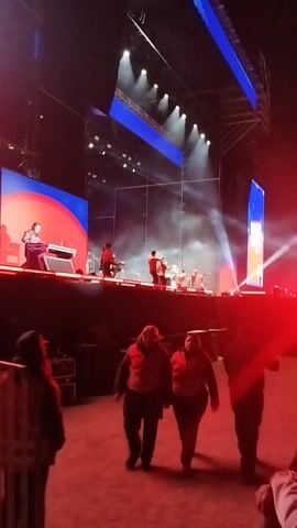 The video captures a live concert or performance at night in an outdoor setting in Maipú, Chile. A band or musical group is performing on a large, brightly lit stage. Several individuals are visible on stage, illuminated by numerous spotlights and stage lights, creating an energetic atmosphere. Large LED screens behind and on the sides of the performers display dynamic blue and red patterns, contributing to the visual spectacle. Haze or smoke effects are present on the stage.

In the foreground, several people are visible, illuminated by a strong red light, suggesting they are either audience members or event staff positioned near the stage. Sound equipment, possibly speakers, is also present in the foreground. The ground in the immediate foreground appears to be a natural surface, possibly dirt or sand. The scene conveys the excitement and vibrancy of a live music event. No discernible text is visible.