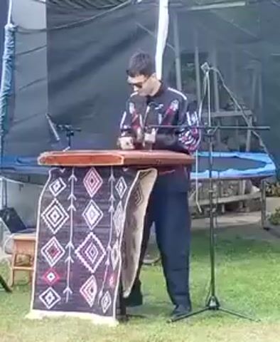 A person in a black jacket with colorful patterns and sunglasses is playing a dulcimer. The dulcimer is on a stand covered by a rug with a diamond pattern. The person is standing and holding two mallets, hitting the strings of the dulcimer. There are microphones set up on stands around the instrument. The scene appears to be outdoors, with grass and a tent-like structure in the background.