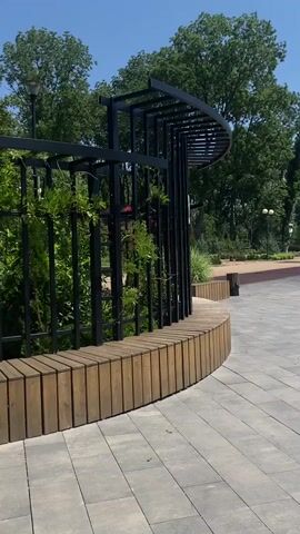 The video shows a park scene on a sunny day with a clear blue sky. In the foreground, a paved area with large, grey rectangular tiles leads towards a curved structure. This structure features a black metal frame with a curved top, resembling a modern pergola, and a bench made of vertical wooden slats. Lush green plants are growing on and around the metal frame. To the right of the bench, the paved area continues, and further back, a pathway with a reddish-brown surface is visible. Along this pathway, there are lampposts and mature trees with dense green foliage. In the middle ground, a small child in a yellow outfit is walking near a water fountain, which is spraying water upwards. To the right of the fountain, another child on a scooter is moving. The background is filled with more trees, and some white structures, possibly tents or small buildings, can be seen. The overall atmosphere is peaceful and suggests a public recreational space.