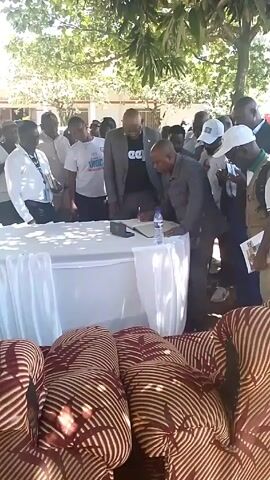 Here's a detailed description of the scene:

**Scene:** The video appears to depict an outdoor event with a formal signing taking place.

**People:**
*   A group of people surrounds a table. Most are standing, observing the signing.
*   One man in a suit is signing a document on the table.
*   Another man in a jacket stands behind him, seemingly overseeing the process.
*   There is a variety of clothing styles, including shirts with logos or writing.

**Objects:**
*   A round table covered with a white cloth is the focal point.
*   A document and a pen are on the table.
*   A water bottle also sits on the table.
*   Patterned cushions or stools are in the foreground.

**Actions:**
*   The primary action is the signing of a document.
*   Other people are observing, possibly taking photos or documenting the event.

**Interactions:**
*   The man signing the document seems to be the focus of attention.
*   The man standing behind him could be providing guidance or overseeing the signing.

**Environment:**
*   The scene is outdoors, with trees and some buildings visible in the background.
*   It appears to be a sunny day.