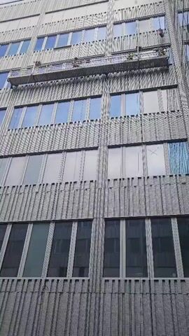 The video shows a low-angle shot of a tall, modern office building with a gray, ribbed facade. Several rows of windows are visible, some reflecting the sky, others appearing darker and possibly covered from the inside.

A suspended scaffolding platform is positioned high up on the building's facade. On this platform, several workers can be seen, appearing to be engaged in maintenance or repair work. They are wearing safety harnesses and are spaced out across the platform. The scene suggests a typical workday with individuals performing tasks at height.

The lighting implies it is daytime, likely overcast or with diffused sunlight, as there are no harsh shadows. The architectural style of the building, with its repetitive window patterns and metallic finish, is characteristic of mid to late 20th-century commercial architecture. The location, Västerås, Sweden, provides a geographical context, suggesting a Northern European urban environment. No specific weather conditions are evident beyond the general lighting. No text is visible in the footage.