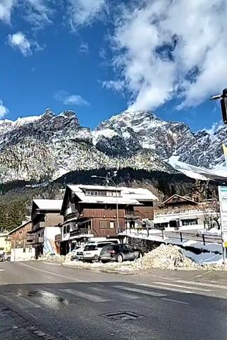 The video showcases a picturesque winter scene in a mountain town, likely in the Alps. The landscape is dominated by snow-capped mountains under a clear blue sky dotted with fluffy white clouds. The foreground features a street with a crosswalk. The street is wet, suggesting recent melting. Several buildings line the street, exhibiting a mix of architectural styles; some are traditional wooden chalet-style structures, while others are modern white buildings. Piles of snow are stacked along the edges of the road and sidewalks, indicating snow removal efforts. A street sign with multiple directions and information is visible, and there are a few parked cars in front of one of the buildings.