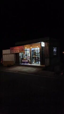 This video shows a series of exterior shots of buildings at night. The first shot is of a small store with its lights on, displaying items inside. The second shot pans to the left, showing several A-frame structures with lights on inside and a tower-like structure in the distance. The third shot shows a white building with a porch and white picket fence, as well as more A-frame structures in the background. The fourth and fifth shots are close-ups of a sign that reads "AWANA" with the words "Glamping Site" below it, and a phone number. The lighting in all shots is artificial and primarily comes from the buildings themselves. The ground appears to be asphalt or a similar dark surface, suggesting a road or pathway. The sky is dark, indicating it is nighttime.
