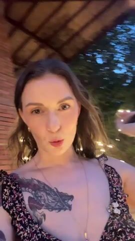 The video shows a close-up shot of a woman with brown hair and a tattoo on her shoulder and chest. She is wearing a floral print dress. The background is blurry but appears to be outdoors at dusk, with trees, lights, and possibly a body of water. There is a structure in the background that resembles the Eiffel Tower. The woman is looking at the camera and her mouth is slightly open as if she is speaking. The video is shaky, indicating it was likely filmed by someone holding the camera while moving.