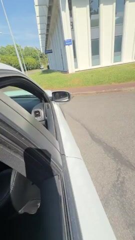 This is a first-person perspective video of someone in a white car with the passenger door open. The car is driving on a paved road with a red curb to the right. There are trees and green bushes lining the road. In the distance, there are buildings and more trees. The sky is blue with some clouds. There is a red and white circular traffic sign visible on the left side of the road. The video shows the interior of the car from the perspective of the passenger seat, with a person in the driver's seat. The car appears to be moving at a moderate speed.