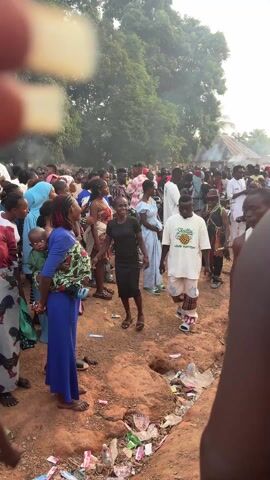The video depicts a large crowd gathered in an outdoor setting, possibly for a celebration or a public event. The ground is dirt, and there's some litter visible. The crowd is diverse, with people of various ages and dressed in a mix of traditional and modern clothing. Some individuals stand out with unique attire, including animal print clothing, beaded necklaces, and headwear. Some people are carrying drums and sticks, suggesting a musical or ceremonial element. The atmosphere appears lively, but the video lacks audio, so it is unclear exactly what is occurring. There are trees in the background and some buildings. The overall impression is a communal gathering with a potential cultural or celebratory focus.