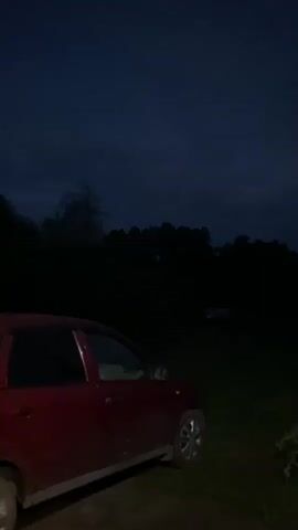 The video shows a dark outdoor scene at night. In the foreground on the left, a red car is parked. In the background, a white car is visible. The sky is dark with some clouds. Trees form a dark silhouette against the sky. The ground appears to be grass and dirt. The scene is dimly lit, possibly by artificial light sources not visible in the frame. There is no movement of people or significant actions depicted.