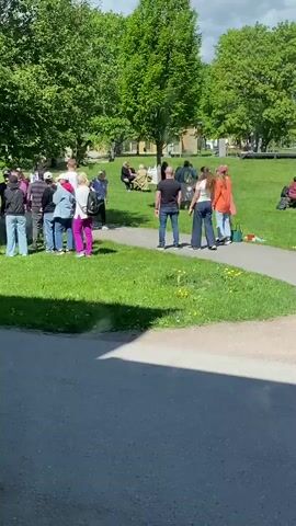 The video depicts a group of approximately 20 people gathered in a park on a sunny day. They are standing on and near a paved path that cuts through a grassy area. Mature green trees surround the area. Some people appear to be watching an activity, while others are sitting or standing nearby. A woman in a blue shirt and black leggings is actively moving, possibly demonstrating or participating in a game or exercise. A few individuals have bags or backpacks. The setting appears to be a public park or green space, possibly hosting a community event or casual gathering. The camera is positioned from an adjacent street.