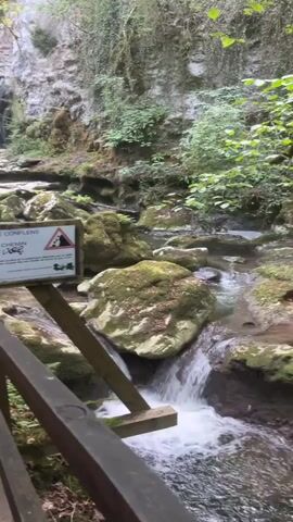 A view of a small waterfall cascading down a rocky cliff face into a stream. A wooden railing runs along the left side of the frame, and a sign with warning symbols is attached to it. The surrounding area is lush with green foliage and moss-covered rocks. The water is clear and flows over smooth stones. The overall scene depicts a natural, serene environment.