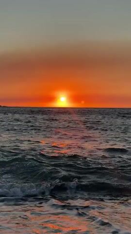 The video shows a sunset over the ocean. The sun is a bright orange disc low on the horizon, casting a reflection on the rippling water. The sky above the sun is a gradient of orange and yellow, fading to a pale blue at the top of the frame. Gentle waves with white foam are breaking on the shore in the foreground, suggesting the viewpoint is from a beach. The overall scene is peaceful and serene.