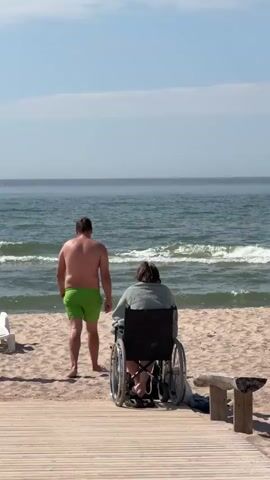 A man in green swim trunks walks away from the camera towards the ocean. A woman in a wheelchair faces the ocean. A woman in a patterned swimsuit walks from the left towards the man. The scene is a sandy beach with a wooden walkway in the foreground and the ocean in the background. The sky is blue with a few clouds.