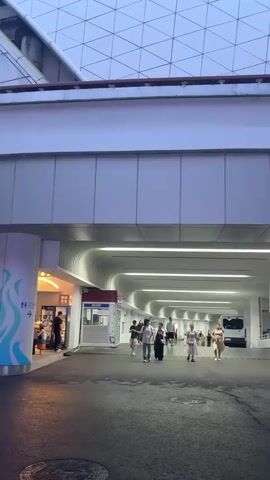 The video shows a wide shot of a street or walkway outside a modern building. People are walking in both directions. The building has a white facade and a unique, curved, illuminated overhang above the walkway. Beyond the walkway, there are palm trees and other greenery. In the background, there's a large screen displaying a person, and tall, industrial-looking structures with platforms. The sky appears to be overcast or twilight. Several individuals are visible, walking alone or in small groups. One person is on a scooter. The overall impression is of a public area in an urban setting, possibly a shopping center or entertainment district.