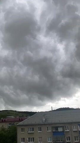 A vertical video shows a dramatic, ominous sky filled with dark, textured clouds that resemble rolling waves or mammatus clouds. Below the sky, the tops of apartment buildings are visible, with some having balconies and white window frames. In the background, a green, tree-covered hill slopes upwards, with some buildings visible on its side. The overall mood is one of impending weather change, possibly a storm.