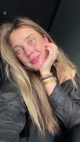 This is a close-up video of a woman with blonde hair wearing a black shirt. She has a nose ring and is wearing several bracelets on her left wrist, as well as a watch. She is resting her chin on her hand and blinking and puckering her lips at the camera. The background is dark and appears to be indoors.