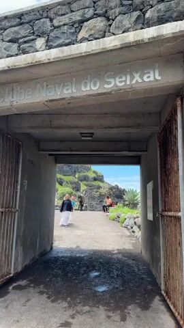 The video shows a stone tunnel leading to a seaside area, likely a recreational or gathering space. The tunnel entrance is marked with the words "Clube Naval do Seixal" in gray lettering above. The tunnel walls are gray concrete, and rusty metal gates are partially open on either side of the entrance. People can be seen walking and sitting beyond the tunnel opening. The area beyond includes rocks, green vegetation, and glimpses of the ocean and a blue sky. One woman is walking toward the tunnel entrance, dressed in black and white clothing. Other individuals are visible in the background, some sitting on the rocks. The pathway through the tunnel appears wet.