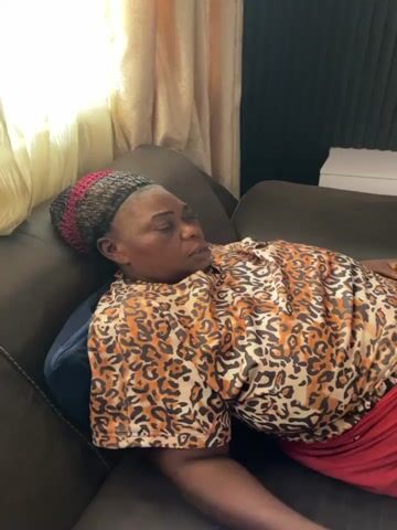 The video captures a woman reclining on a brown sofa in an indoor setting, likely a living room. She is wearing a short-sleeved, leopard-print top, a red skirt or wrapper, and a multi-colored knitted hat (black, red, and grey). A blue pillow is placed under her head.

At the beginning of the clip (0:00), the woman is lying down with a neutral expression, looking towards the left side of the frame. Natural light streams in from a window on the left, partially covered by light-colored curtains. From 0:02, she lifts a light green mug to her mouth and takes a sip. Another hand briefly appears at the bottom left, holding a blue-capped plastic water bottle. The background includes a dark wall and a white shelf on which a phone is placed. The woman appears relaxed and calm. The scene suggests a comfortable domestic environment in Jos, Nigeria. No visible text is present in the video.