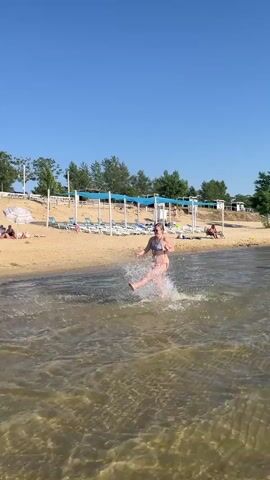 A woman in a bikini is playfully splashing through the water at a beach. She is mid-stride, with water spraying around her legs. The sun is shining brightly, indicating it is daytime, and the sky is clear blue. In the background, a sandy beach is lined with lounge chairs and umbrellas, suggesting a resort or public beach area. A few other people can be seen on the beach, some sitting and others standing. Trees are visible further back, and a blue canopy structure stretches along the shoreline. The overall atmosphere appears relaxed and enjoyable, with the woman's action conveying a sense of fun and freedom. There is no visible text in the media.