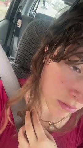 The video shows a close-up, first-person perspective from inside a car. The focus is on the person's face and upper body, with their hand touching their neck. The person is wearing a red shirt and a necklace. The car's interior is visible, including the seatbelt and the car seat. The background is blurred, but suggests an outdoor environment, possibly a street. The person's hair is brown and somewhat disheveled. The lighting suggests daylight.