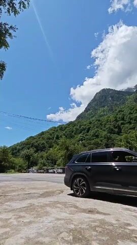 The video opens with a side view of a black SUV car parked on a paved surface. The vehicle has dark tinted windows and distinctive multi-spoke wheels. In the background, lush green mountains rise under a bright blue sky dotted with fluffy white clouds. A sign with some text, possibly "CAFE COJIKA", is partially visible on the left side near the car.

The camera then pans slowly to the right, revealing more of the scenic mountainous landscape. The mountains are densely covered with trees, primarily evergreens and deciduous trees with vibrant green leaves. A paved road stretches across the mid-ground, and a large puddle reflects the sky on the rough concrete surface in the foreground. Further along the road, several small wooden structures or gazebos are visible amidst the trees. The overall scene depicts a sunny day in a picturesque, mountainous region.