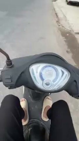 The video shows a first-person perspective of someone riding a scooter on a road. The rider is wearing black pants and beige slides. The scooter is black with a digital display. The road has white lines marking lanes. The environment is a town with buildings and trees visible in the background.