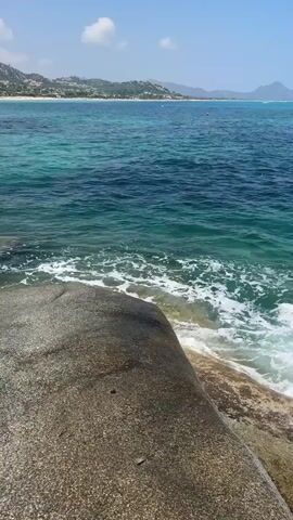 The video shows a body of water with waves breaking against a large rock in the foreground. In the background, there is a coastline with buildings and hills under a clear blue sky with scattered clouds. The water is a deep blue transitioning to turquoise closer to the shore. The waves are white and foamy as they crash against the rock. The sun is shining brightly, casting shadows and illuminating the scene. The overall impression is a serene and beautiful coastal landscape.