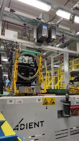 A woman wearing a navy blue t-shirt, black pants, and grey work gloves is standing at a factory assembly line. She is facing away from the camera and appears to be working on a piece of machinery mounted on a conveyor. The machinery is yellow and silver and has a circular opening in the center. The woman is using her hands to manipulate something within the circular opening. The environment appears to be a brightly lit factory with various machines and equipment visible in the background, including overhead lights and pipes. There are also warning signs with hazard symbols visible on the machinery and conveyor. The woman's hair is pulled back in a ponytail.