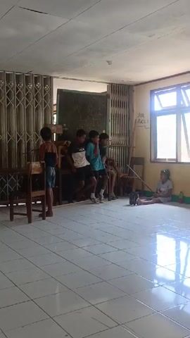 A group of children are in a room with tiled floors. Some children are sitting on a bench, while others are standing. One child is sitting on the floor. The room appears to be a classroom or a common area. There are windows, doors, and a blackboard visible. The children are casually dressed. Some are looking towards the camera. One child is holding a drink.
