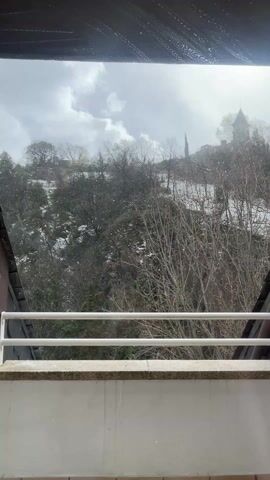 The video shows a view from a balcony on a snowy day. The balcony has a white railing and a light-colored stone surface. Beyond the railing is a hillside covered with bare trees and patches of snow. A building with a tall steeple or tower sits at the top of the hill. Snowflakes are falling and visible against the overcast sky. The video is taken from indoors, and the top of the frame shows the edge of a dark overhang or roof.