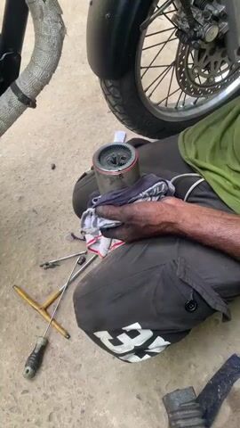 This video shows a person's hands, which appear to be dirty, holding a metal cylindrical object with gears inside. The person is wiping the object with a cloth. There are tools, including a screwdriver and a wrench, lying on the ground near the person. Part of a motorcycle wheel and exhaust pipe are also visible in the background. The ground appears to be dirt or gravel.