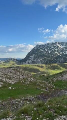 The video presents a scenic view of a mountainous landscape on a sunny day with a blue sky and scattered white clouds. The foreground is composed of a rocky terrain with patches of green grass. In the midground, there are rolling hills and valleys. The background showcases majestic mountains, some with snow-capped peaks. The camera pans slightly to the right, revealing different angles of the mountain range.  No people or other objects are visible in the video.