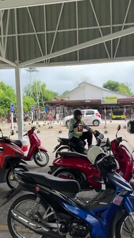 Under a large, metal-framed canopy, several motorcycles are parked in a row. The motorcycles are a mix of colors: red, blue, black, and white. A person wearing a helmet and a reflective vest with the number 19 is seated on one of the motorcycles. In the background, buildings and greenery are visible. The sky is overcast.