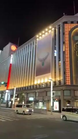 The video showcases a nighttime street scene in what appears to be a bustling urban area. The camera pans across several large buildings, highlighting their architectural styles and prominent advertisements. One building features a large Bulgari advertisement. Streetlights illuminate the scene, and multiple cars, mostly white vans, are seen moving along the road. The environment appears to be modern and well-lit, possibly a commercial district.
