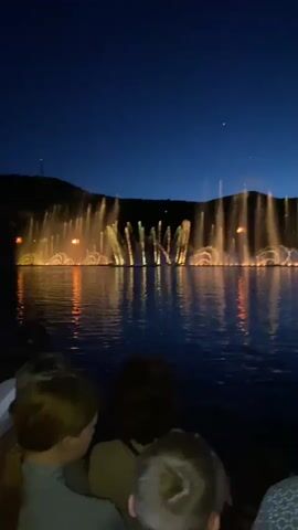 This is a short video clip of a fountain show at night. The fountain is illuminated with bright lights that change color from orange to red. The water jets are spraying into the air and reflecting the light, creating a dazzling effect. In the foreground, the backs of several people's heads are visible as they watch the show. The sky is dark blue, indicating nighttime, and a faint outline of hills can be seen in the background. The water in the foreground is calm, with ripples reflecting the lights of the fountain.