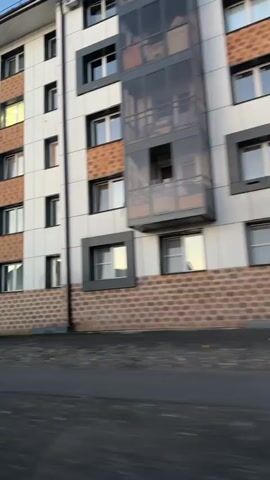 A side view of a modern apartment building with a white facade and brown brick accents. Several windows and balconies are visible. A white SUV is parked on the left side of the frame. A yellow traffic cone is visible in the foreground. The camera is moving, suggesting it is mounted on a vehicle. The lighting indicates it is either early morning or late afternoon.