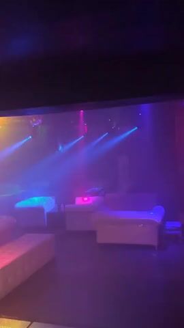 The video pans across a dimly lit, enclosed space that appears to be a nightclub or lounge. White tufted couches and benches are arranged in small seating areas. The environment is filled with colored stage lighting, primarily blue, green, and purple, with some pink and yellow hues also present. Smoke or haze is visible, catching the light beams. In the background, there is a stage area with lighting fixtures and what looks like a DJ booth. A single person, wearing dark clothing, is visible standing on the left side of the stage, appearing to be observing the room. There are no overt interactions or activities taking place, and the overall impression is one of an empty venue preparing for or between events.