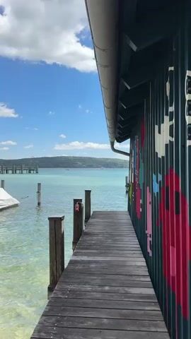 A wooden pier leads to a building with colorful graffiti on its side, adjacent to a clear, turquoise body of water under a blue sky with white clouds. In the distance, a tree-covered landmass is visible, with a pier extending into the water. The camera is positioned on the pier, looking towards the water and the building.