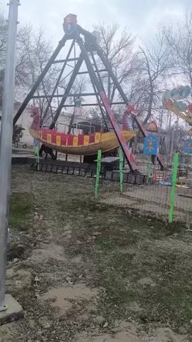 The camera films a small, fenced-off amusement park, revealing two stationary rides. One ride resembles a Viking ship with a dragon head, painted in yellow, red, and black, resting on a black frame. A spinning swing ride with colorful swings sits to the right of the ship ride. Bare trees surround the park. The ground is a mix of dirt, mud, and sparse grass. The sky is overcast. Hindi music plays in the background. The camera remains still for the duration of the video.