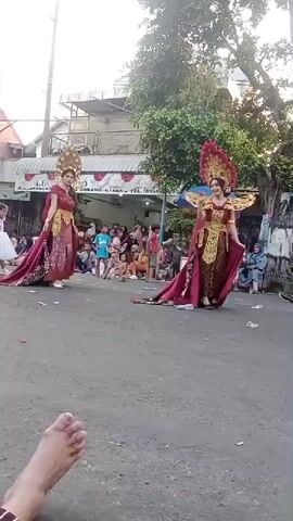 The video shows a street parade or performance with several participants in elaborate costumes. Two women in striking red and gold traditional dresses with ornate headpieces are the central focus. They are positioned on a paved street, likely a road cleared for the event, with a crowd of spectators gathered on the sides. The spectators are seated or standing, watching the performance. In the background, there are buildings, trees, and street signage, suggesting an urban or semi-urban setting. A child in a white dress also appears in the scene, possibly part of the performance or a spectator. The lighting indicates it is daytime, likely late afternoon or early evening. One shot includes a close-up of a person's feet in the foreground, indicating the perspective of someone sitting or lying down to watch.